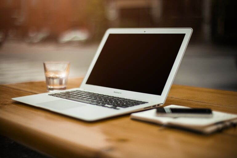 Laptop, glass and a notebook and pen on a wooden desk with a blurred background