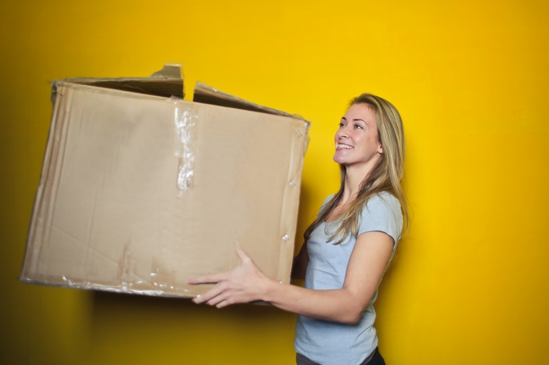 Smiling woman carrying a large cardboard box against a yellow background
