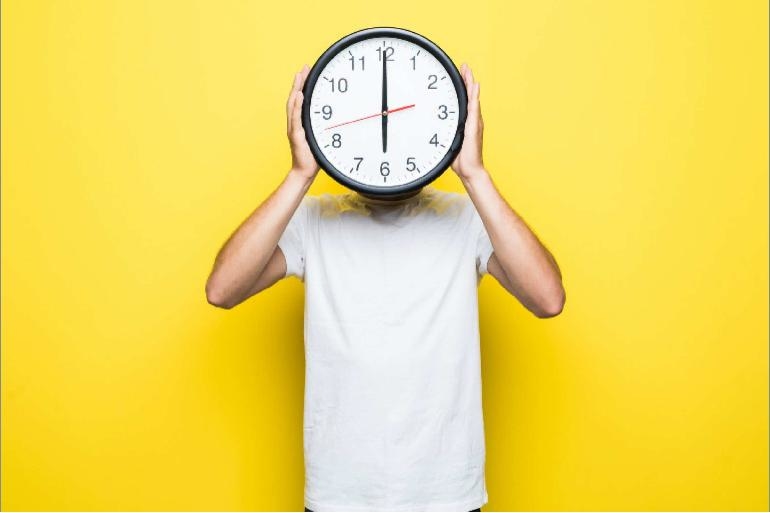 Person in a white t-shirt against a yellow background holding a clock in front of her face
