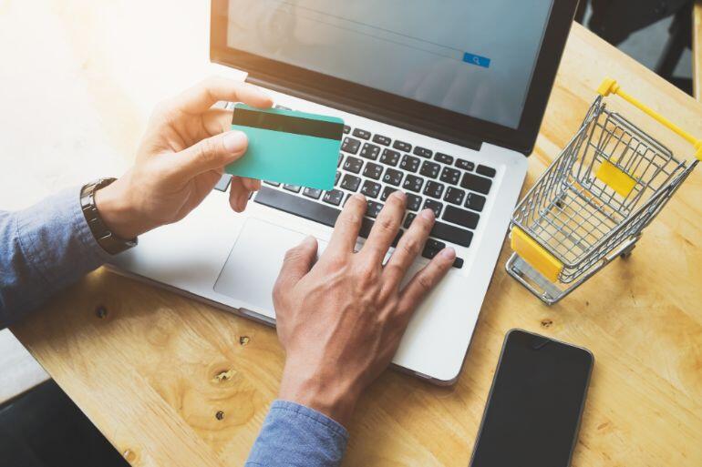 Person holding a credit card over a laptop keyboard with a small shopping cart next to it