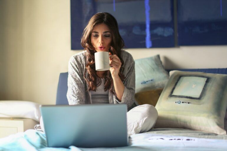 Woman sitting on a bed, in front of a laptop drinking a coffee
